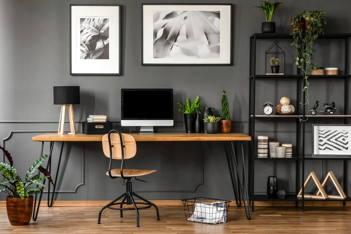Stylish home office with a wooden desk, computer, and chair. Gray wall with black-and-white leaf art. Shelves hold plants and decor for a modern vibe.