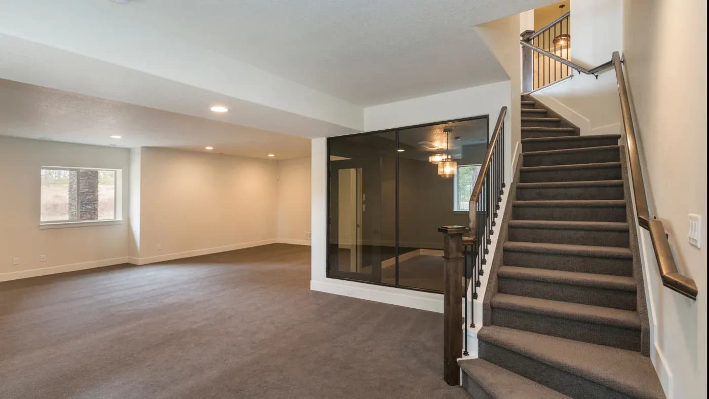 Spacious empty room with beige walls and brown carpet, featuring a large window, recessed lighting, and dark wooden staircase on the right. Cozy ambiance.