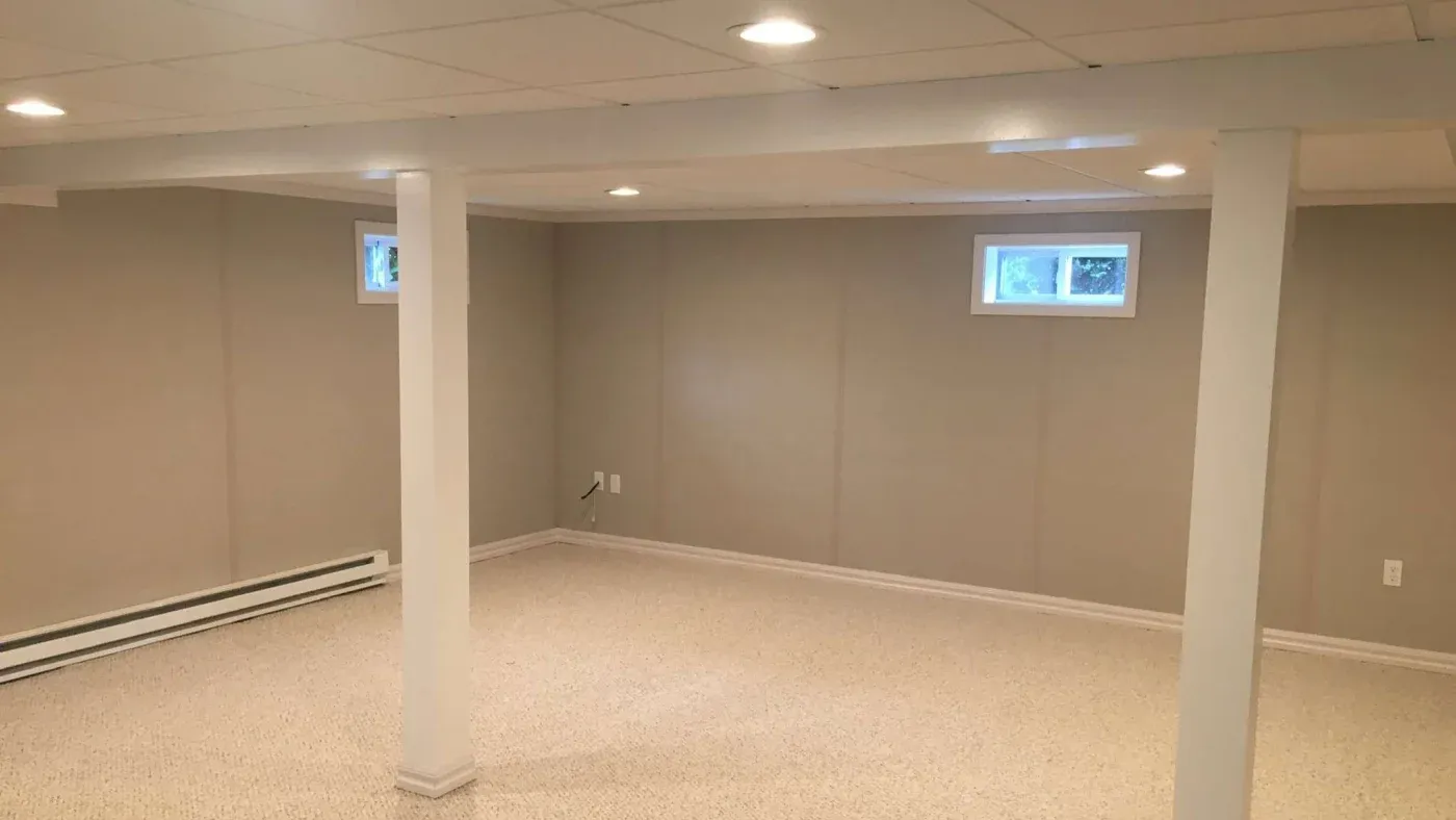 A spacious, empty basement with beige walls, carpeted floors, recessed lighting, and small rectangular windows, creating a clean, neutral atmosphere.