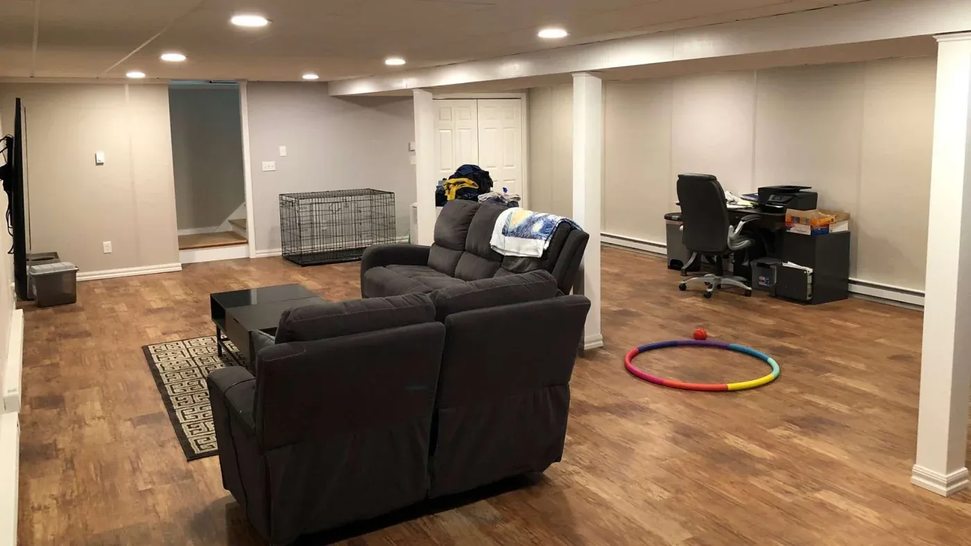Spacious basement with wooden floors, gray sectional sofa, small coffee table, office desk, and chair. Dog crate, colorful hoop, and toys on floor.