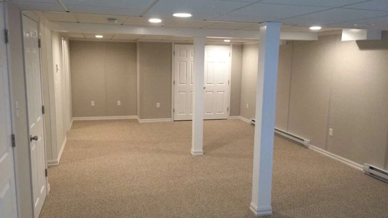 A spacious, empty basement room with beige carpet and light grey walls. It features white support beams and recessed ceiling lights, conveying a clean, unfinished look.