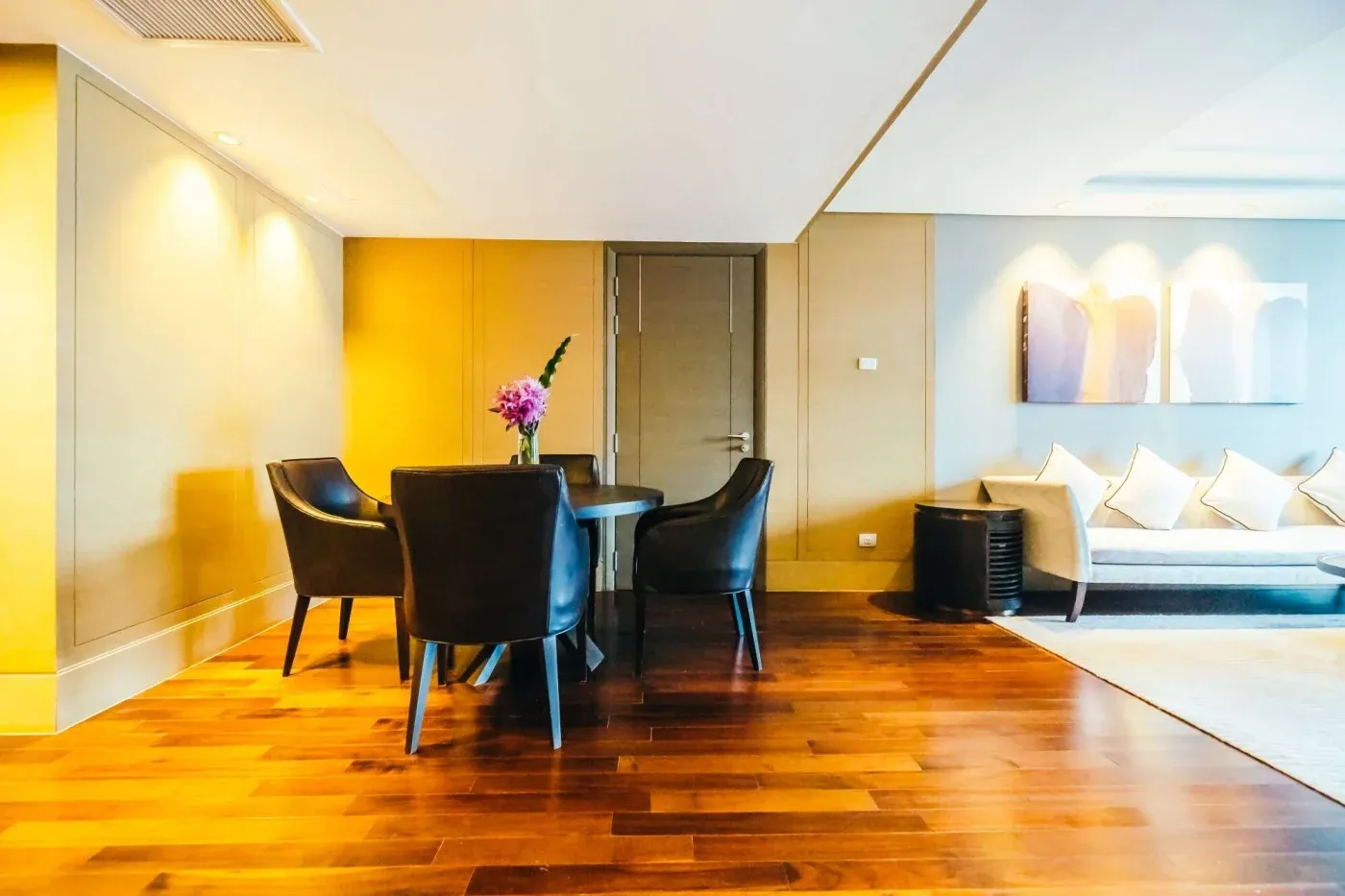 A modern living room with wood flooring, featuring a cozy sitting area with a three-seater sofa and cushions. A round table with black chairs and a vase of purple flowers sits beneath warm lighting, creating an inviting atmosphere.