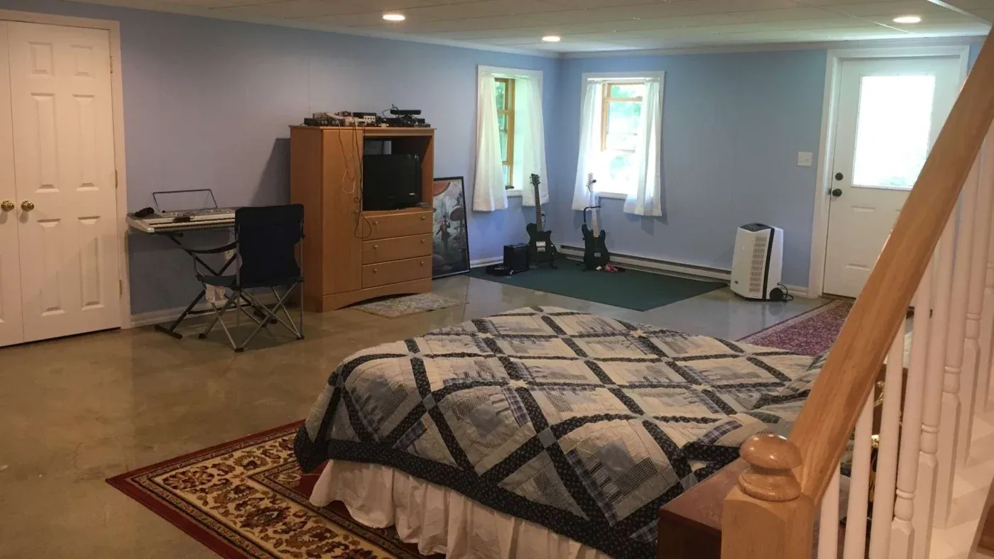 A spacious basement room with light blue walls features a bed with a geometric quilt, a wooden dresser with a TV, two windows, and a small desk with a folding chair.