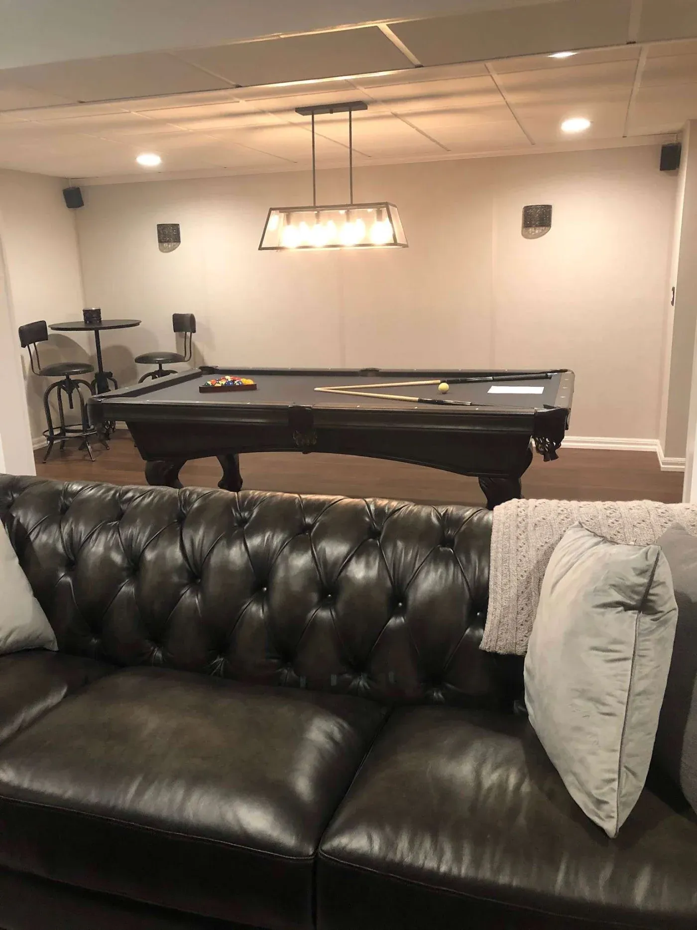 A cozy room features a tufted leather sofa with cushions and a throw blanket in the foreground. Behind is a pool table with cue sticks and balls, under a modern chandelier. A small round table with two barstools is in the corner, creating an inviting atmosphere.