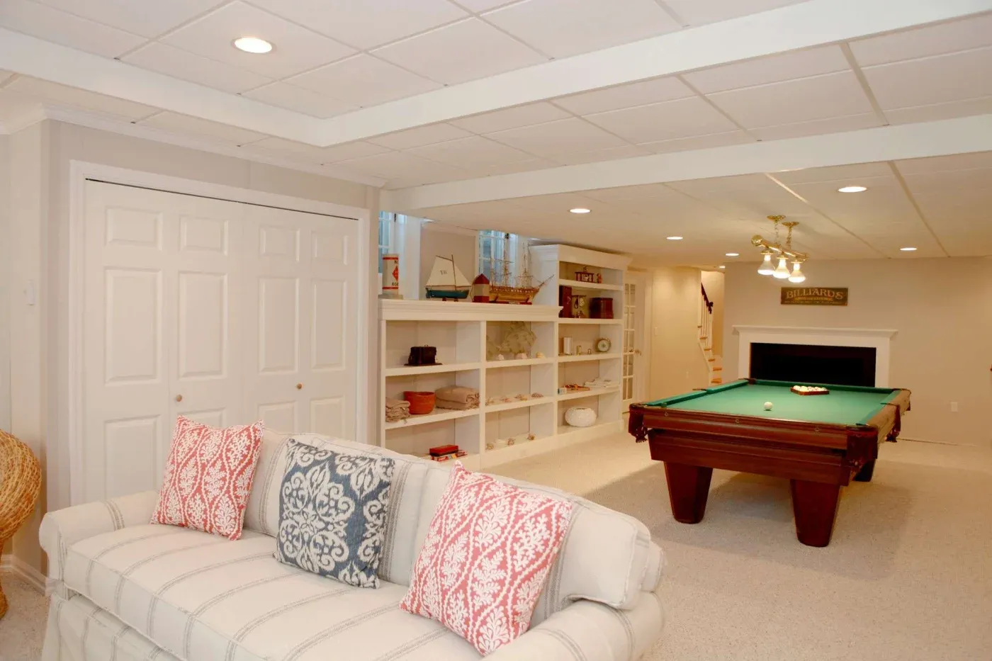 A cozy basement room with a pool table under soft lighting, white shelving filled with decor, and a sofa with red and blue patterned pillows.