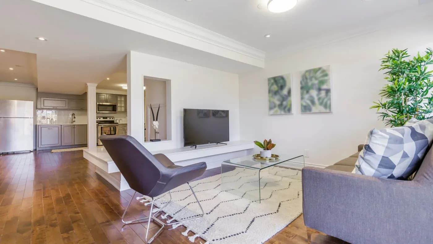 Modern living room with a gray sofa, geometric pillow, black chair, glass coffee table, and TV. Bright ambiance and wooden floors add warmth.