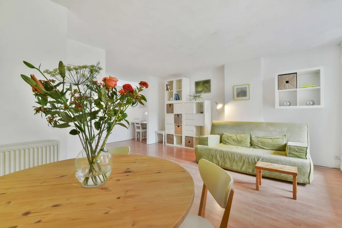 Bright living room with a wooden table and orange flowers in a vase. Green sofa, wooden floor, white shelving, and soft lighting create a cozy atmosphere.