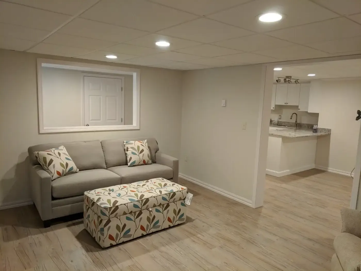 A cozy basement room with a gray sofa and patterned ottoman, featuring colorful leaf designs. Adjacent is an open, bright kitchen. The space feels warm and inviting.