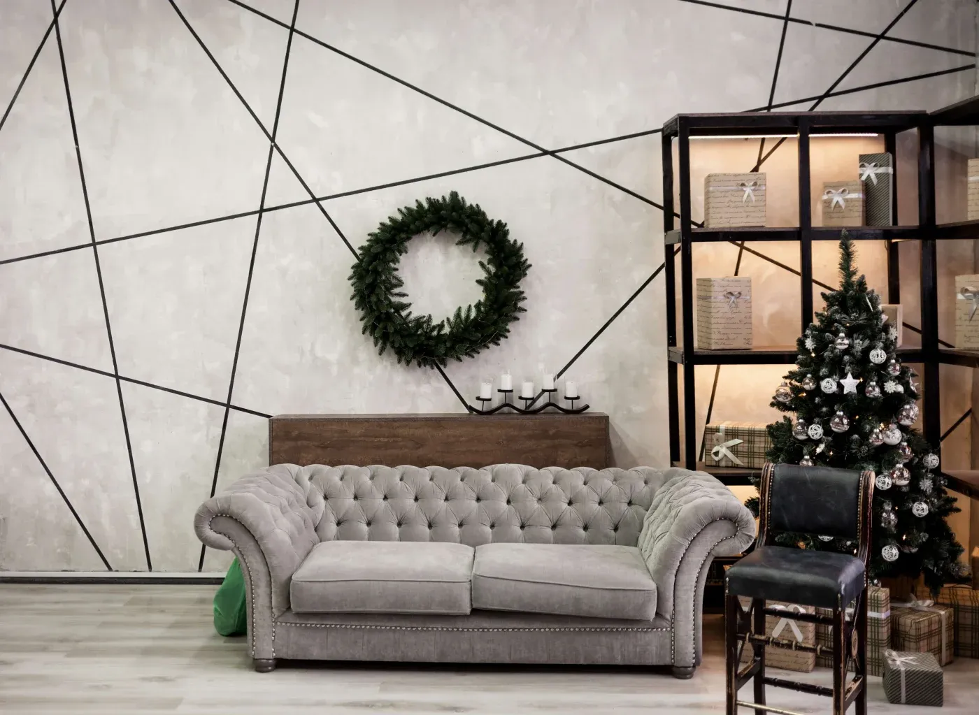 Modern living room with a gray tufted sofa, geometric wall design, and a decorative wreath. A small, decorated Christmas tree and gift boxes add festivity.