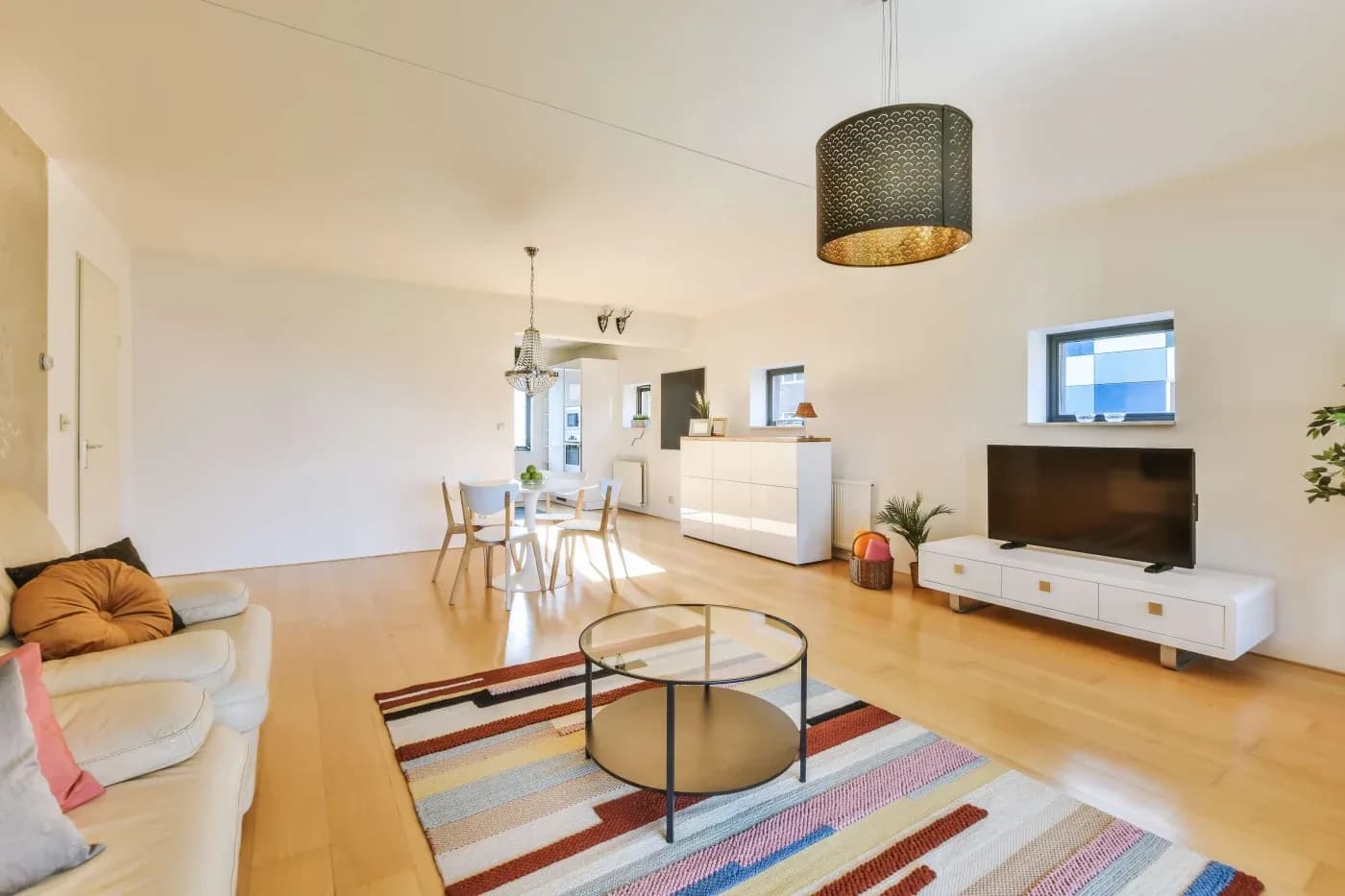 Modern living room with light wood flooring, featuring a cozy beige sofa, circular glass coffee table on a colorful striped rug, and a flat-screen TV on a white stand. A dining table with chairs and pendant lights are visible in the background, creating a bright and inviting atmosphere.