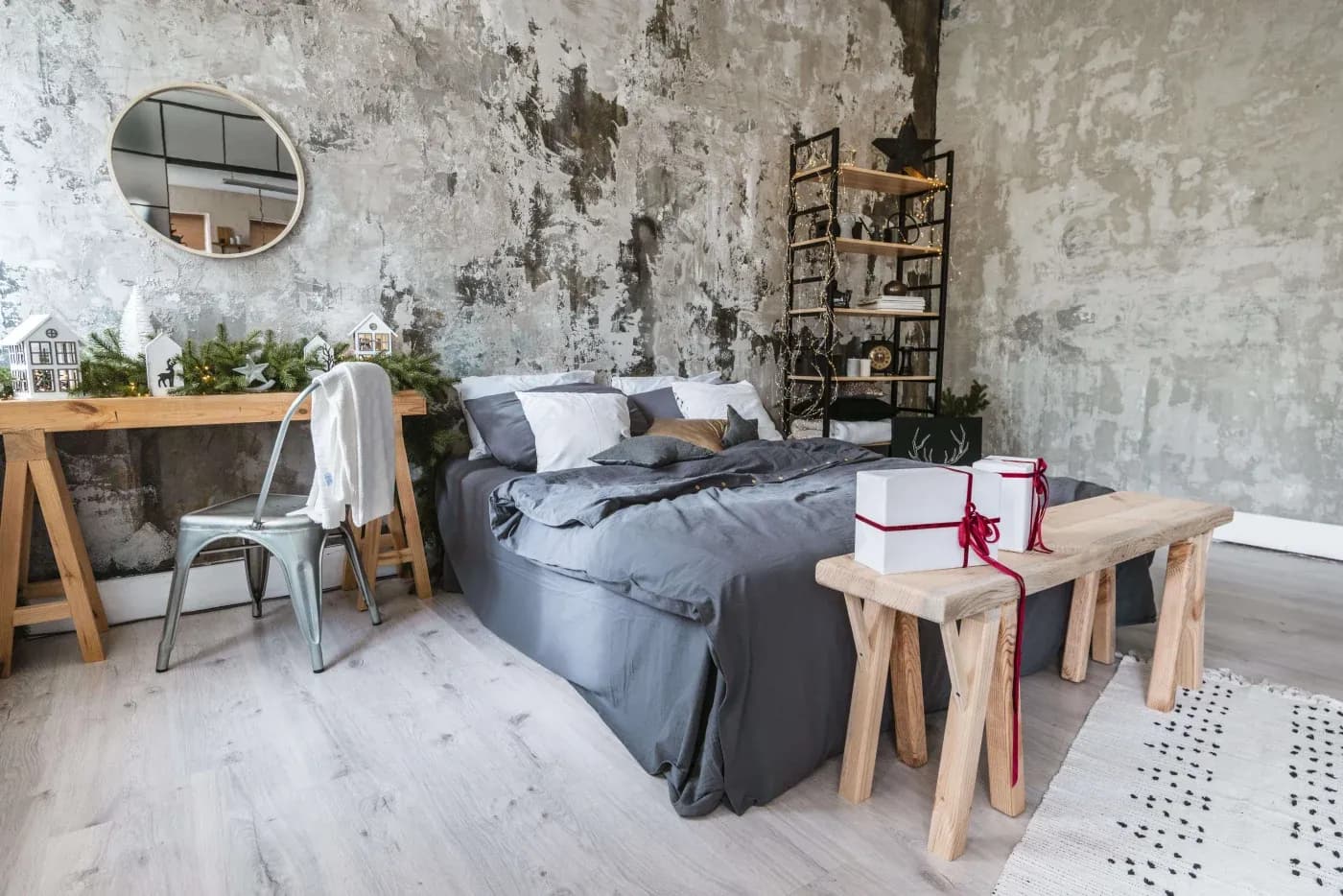 A cozy, modern bedroom with a textured gray wall, gray bedding, a wooden bench with gift boxes, a rustic desk with decor, and soft lighting enhance a relaxing ambiance.
