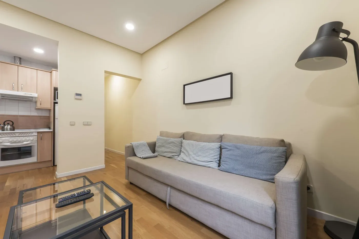 Modern living room with a beige sofa, striped cushions, and a minimalistic lamp. Adjacent kitchen with wooden cabinets. Cozy and inviting ambiance.