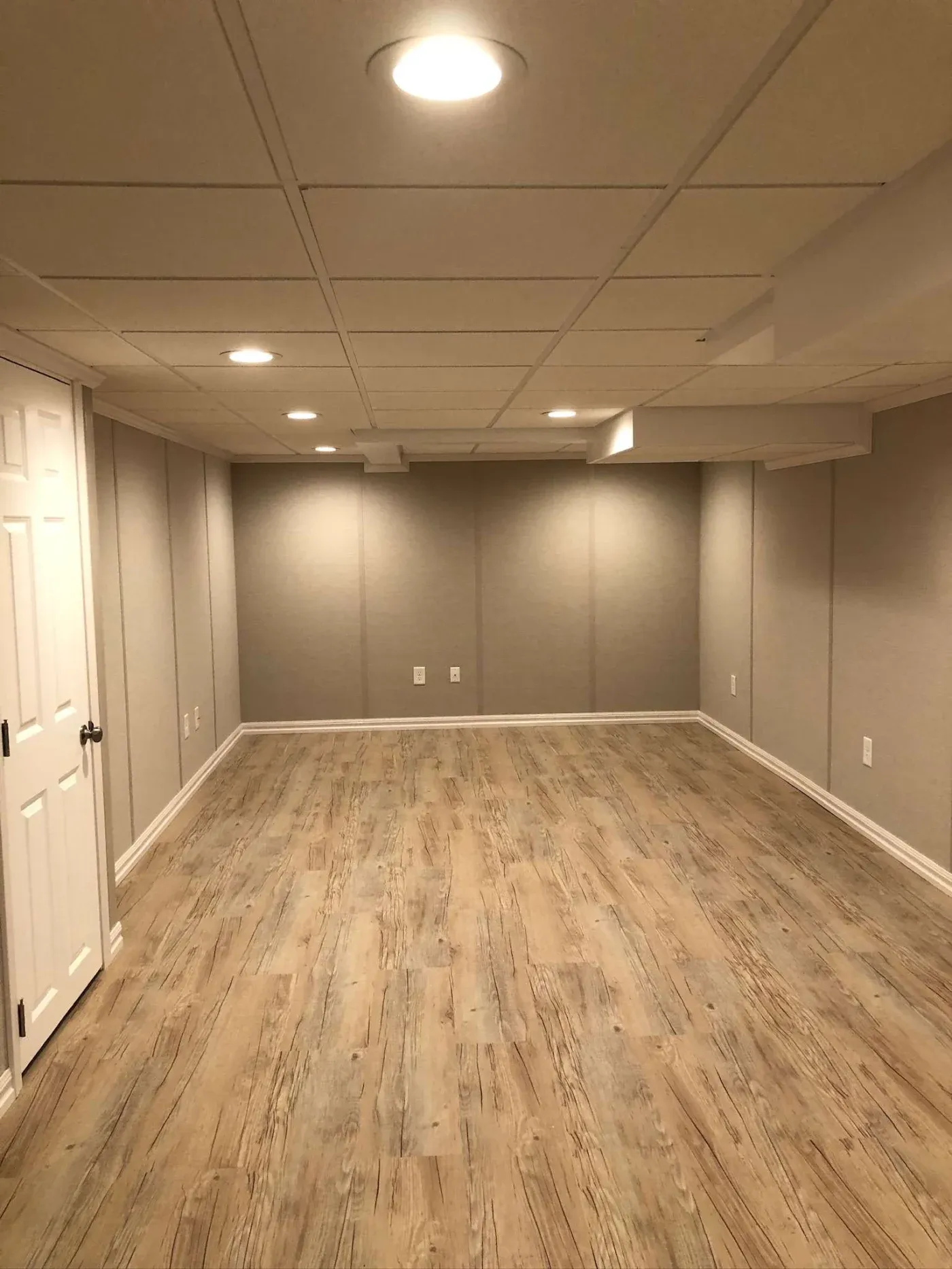 A newly renovated basement with light wood flooring, beige walls, and a white grid ceiling. Soft lighting from recessed fixtures creates a cozy ambiance.