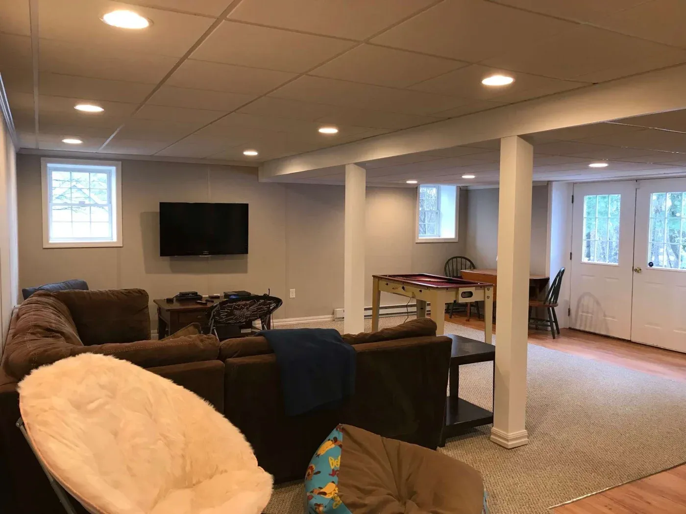 Spacious basement with cozy brown sofas, a wall-mounted TV, and a plush cream chair. A small table and chairs are near a window, creating a relaxed ambiance.