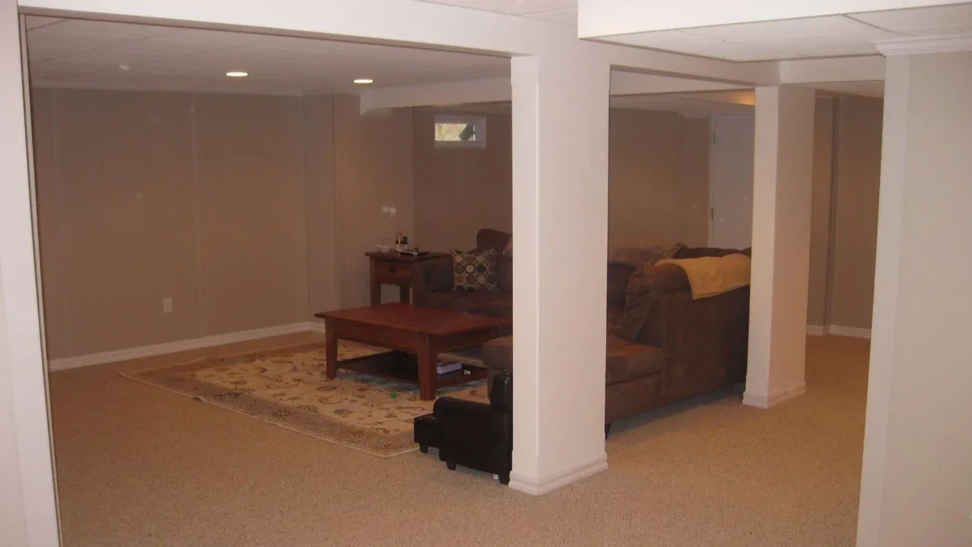 Spacious basement with beige walls and carpet, featuring a wooden coffee table on an area rug, surrounded by a brown sectional sofa and soft lighting.