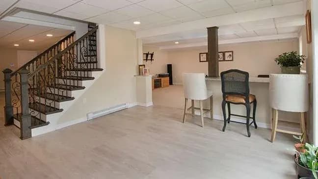 Spacious basement with light wood flooring, a staircase with ornate railing, a bar area with three chairs, and a potted plant, creating a cozy atmosphere.