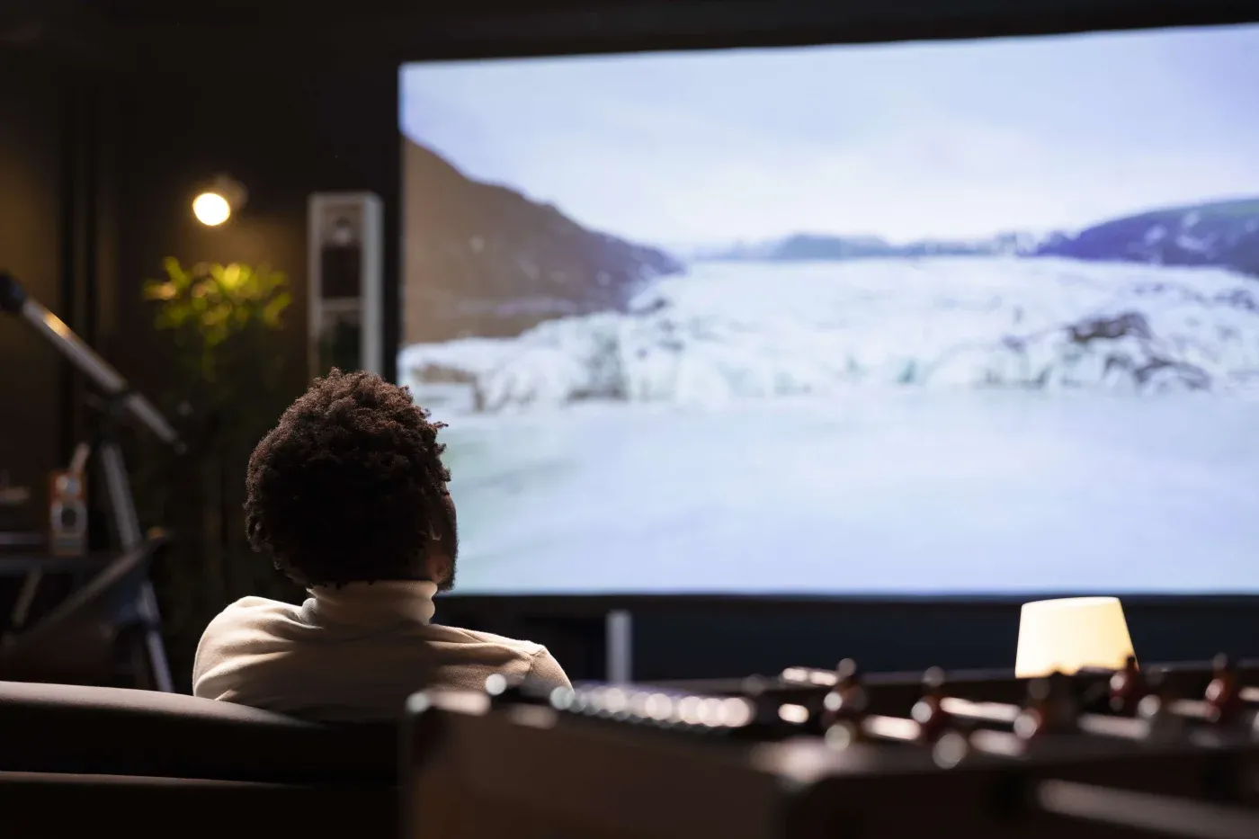 A person seated in a dimly lit room watching a large screen displaying a snowy landscape. The atmosphere is cozy and focused, with a warm lamp nearby.