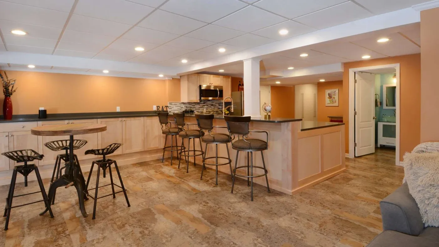 A cozy basement bar with light wood cabinets, stone countertops, and five metal bar stools. A round table with stools sits on a textured floor. Warm lighting and a homey vibe.