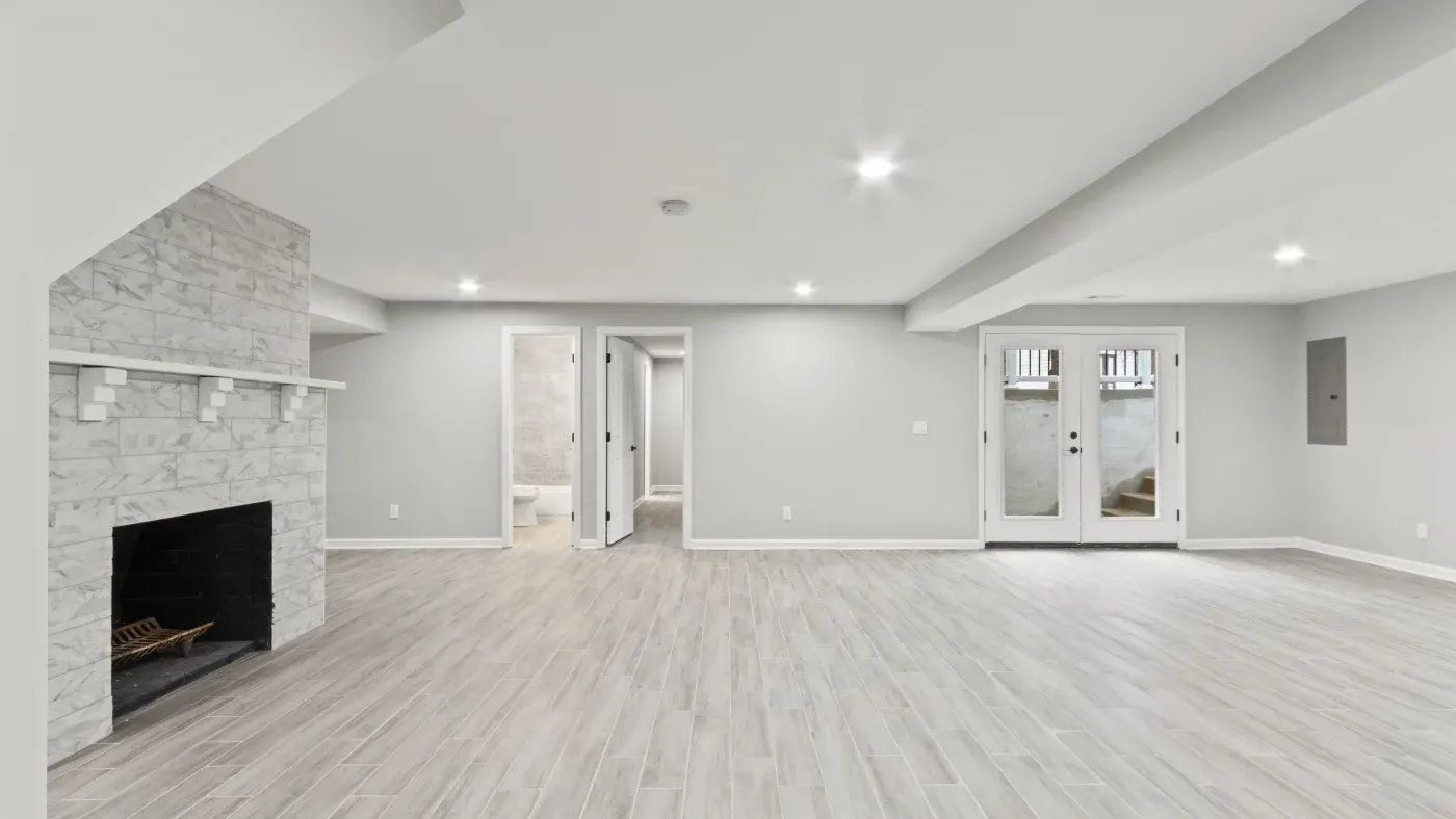 A spacious, bright basement with light gray flooring, white walls, and a stone fireplace. French doors and recessed lighting enhance the airy feel.