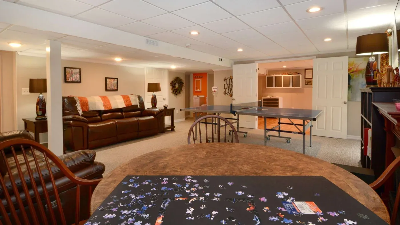 A cozy basement with beige walls features a brown leather sofa, a round table with a jigsaw puzzle, a ping-pong table, and warm lighting, creating a welcoming atmosphere.