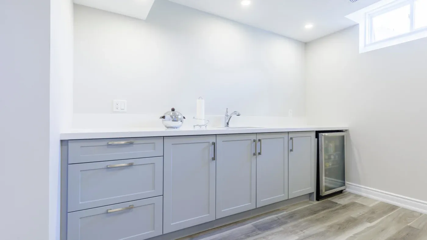 Modern kitchenette with light gray cabinets, sleek countertop, small sink, and mini fridge. Soft lighting and light-colored wooden floor convey a calm, clean ambiance.