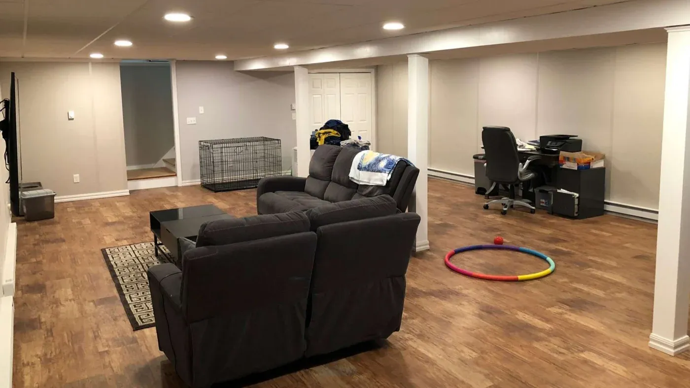 A spacious basement with hardwood floors features a gray couch, coffee table, and rug. A dog crate, office desk, and colorful hula hoop are visible.