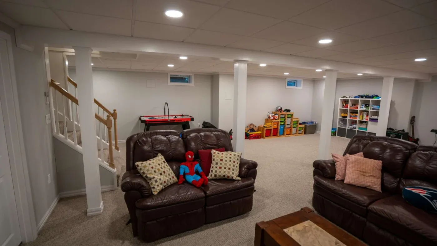Cozy basement with brown leather sofas, a Spider-Man toy, and patterned pillows. Air hockey table and organized toys create a playful, inviting atmosphere.