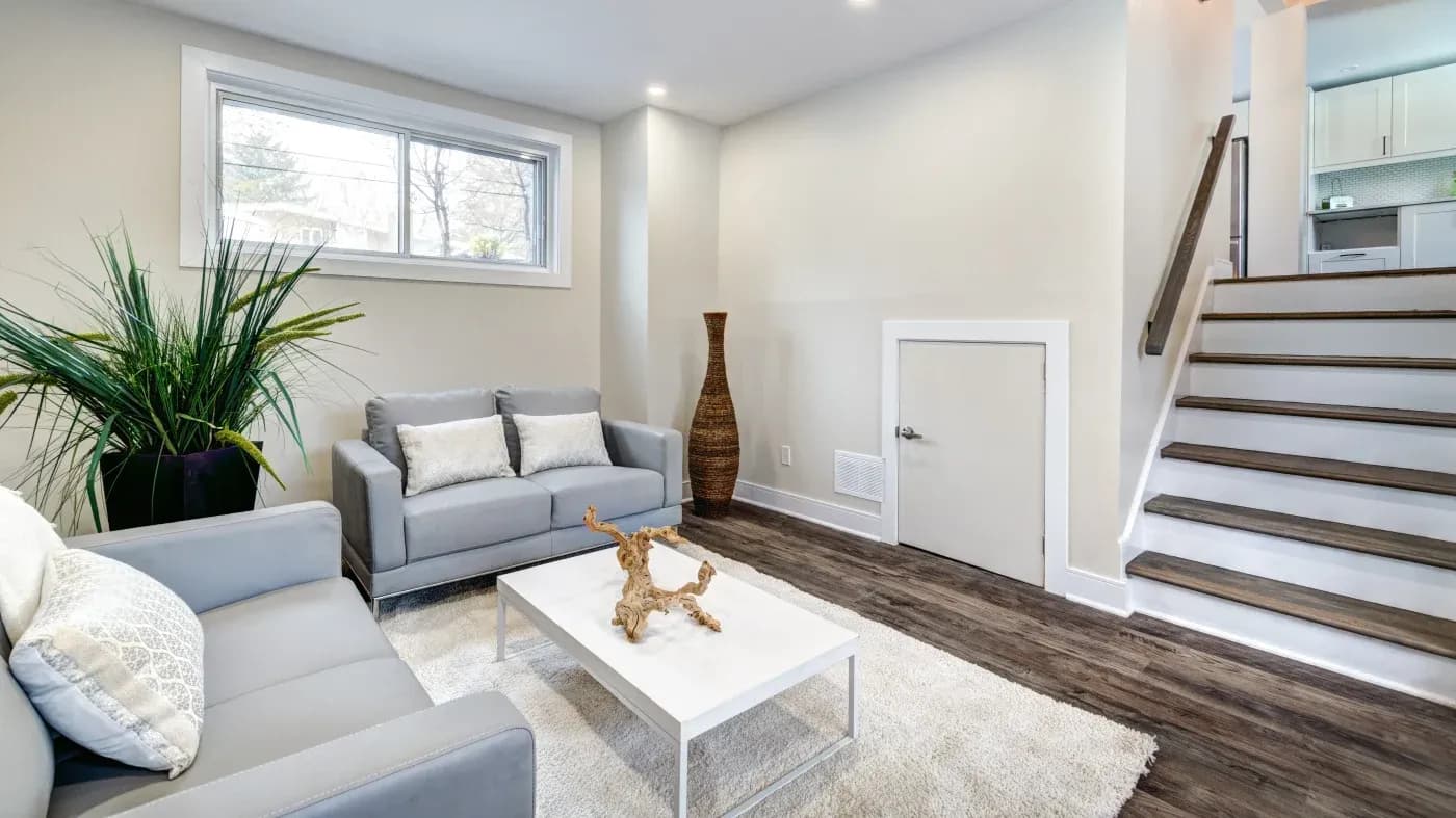 Modern living room with gray sofas, white cushions, and a central white coffee table featuring a decorative branch. A large plant and vase add greenery. Steps lead to a kitchen area. Neutral tones and natural light create a bright, welcoming atmosphere.