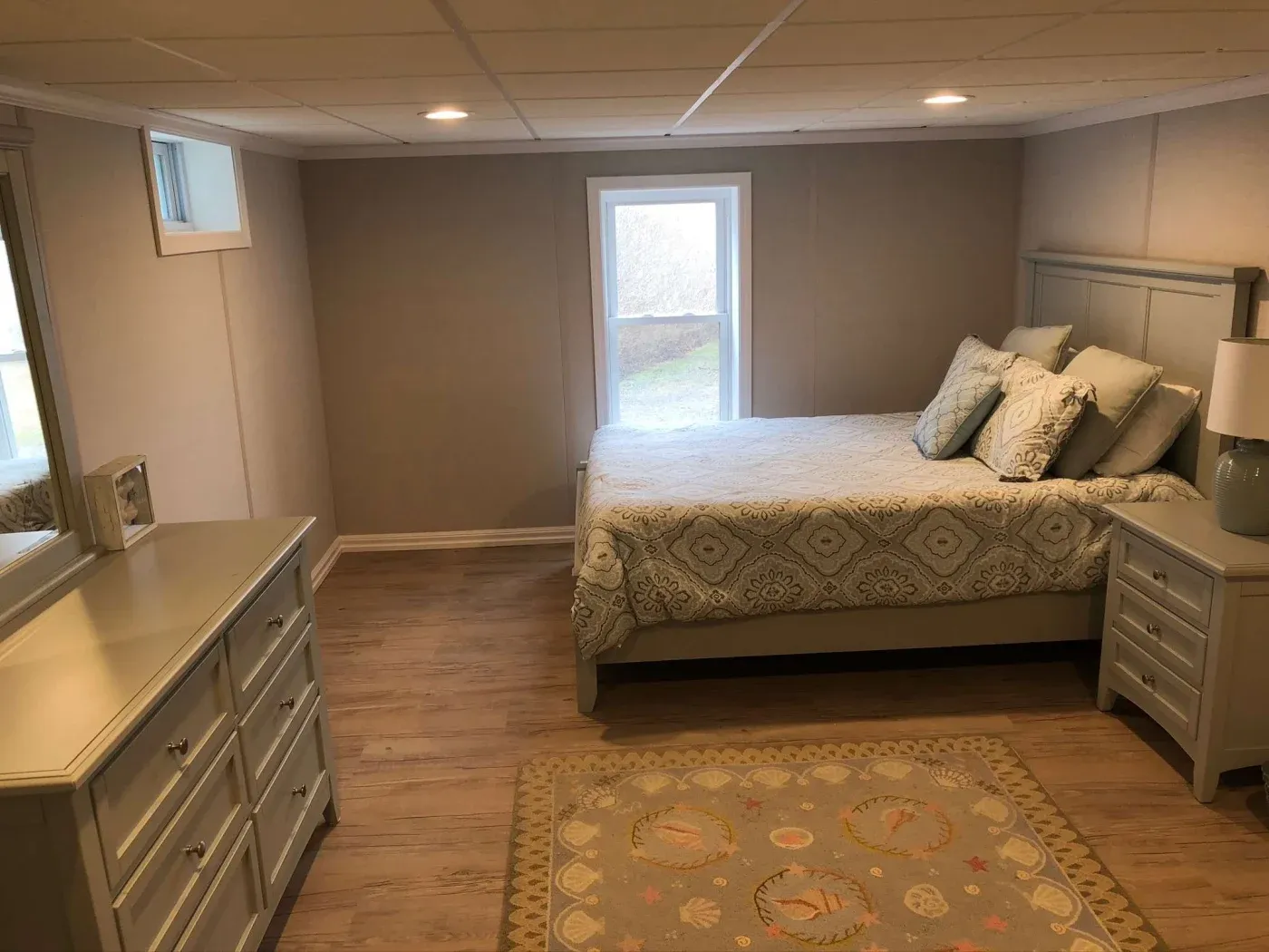 Bright bedroom with a queen bed, patterned bedding, and pillows. A window lets in natural light. There's a gray dresser and matching nightstand with a lamp.