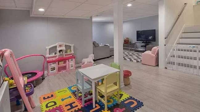 A cozy basement playroom features colorful toys, a white child-sized table set on an alphabet mat, with a sofa and TV in the background. Bright and inviting.