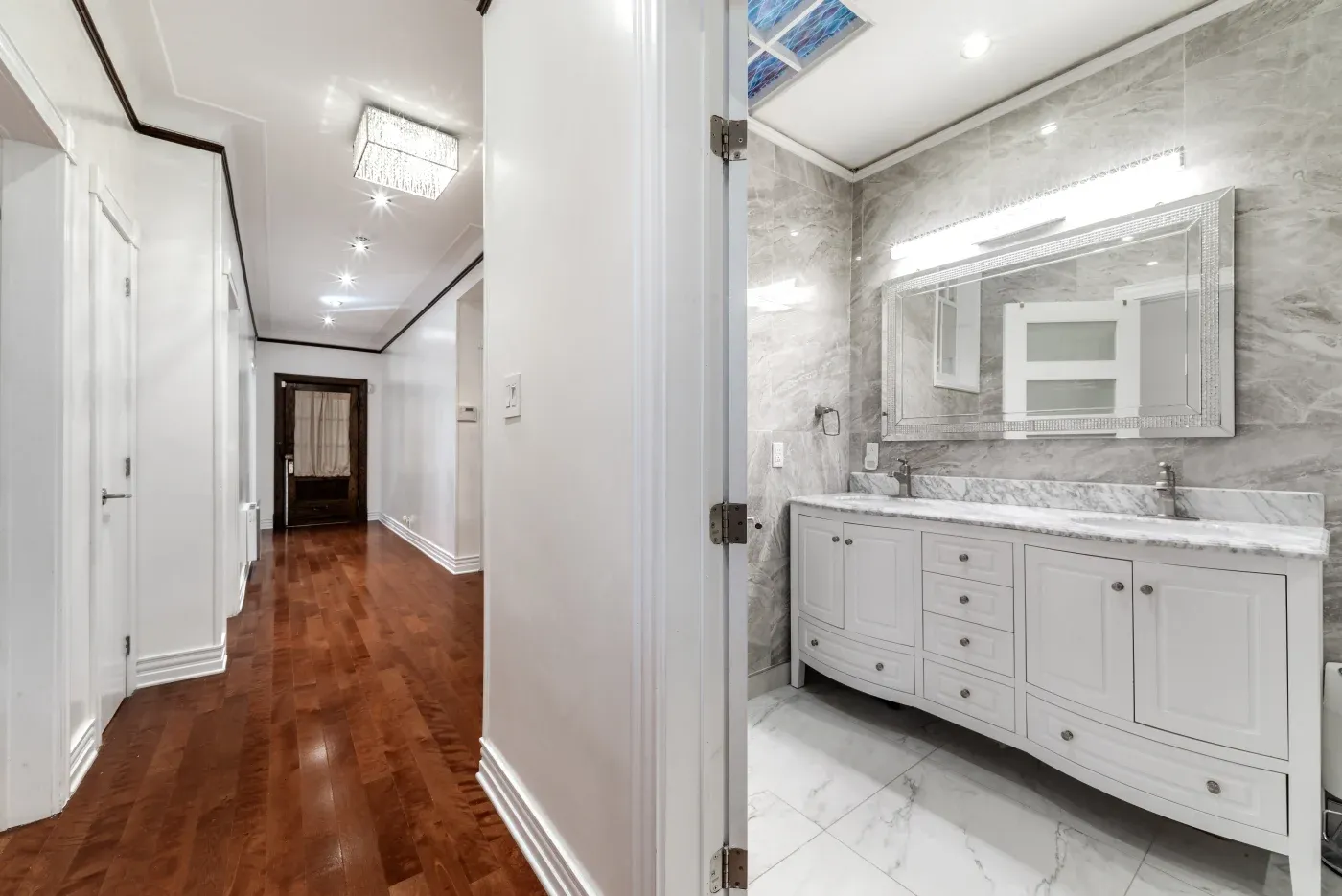 Bright hallway with polished wooden floors and a crystal chandelier. Open door reveals a modern bathroom with marble tiles and a sleek double vanity.