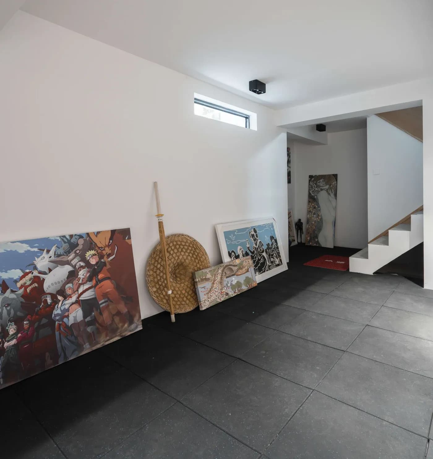 Interior with a white wall and black tiled floor showcasing several anime and art prints leaning against the wall. A staircase is visible on the right.
