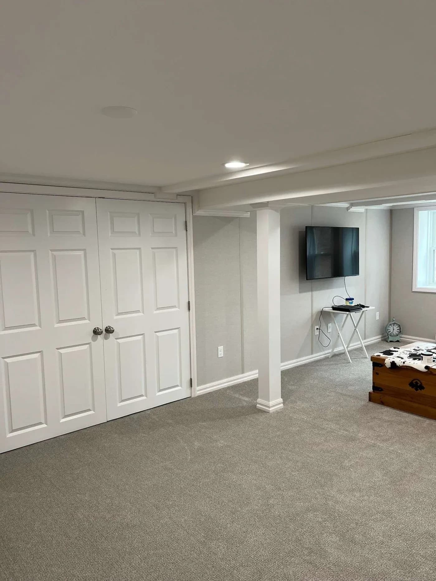 A clean, well-lit basement with light gray carpet. Closed white double doors on the left, a wall-mounted TV, and wooden chest on the right create a cozy, organized atmosphere.