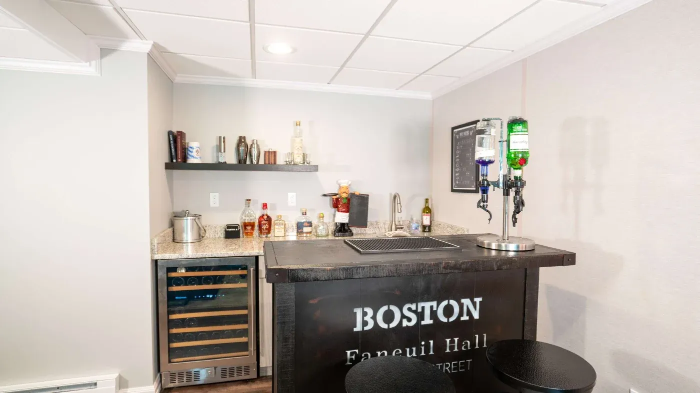 A cozy home bar with a black counter labeled "Boston Faneuil Hall." It features liquor dispensers and shelves with bottles. The atmosphere is inviting.