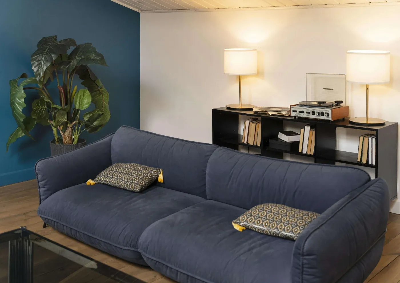 Modern living room with a plush blue sofa, patterned cushions, a record player on a black shelf, books, and warm lighting; a relaxed, cozy ambiance.