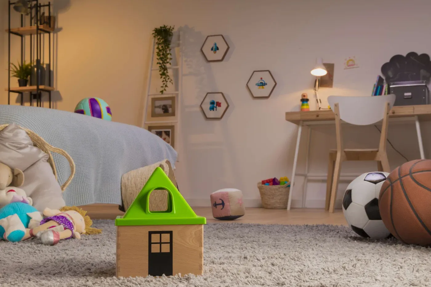 A cozy child's room with toys strewn on a carpet, including a wooden block house, dolls, and soccer and basketballs. A desk and bed are in the background.