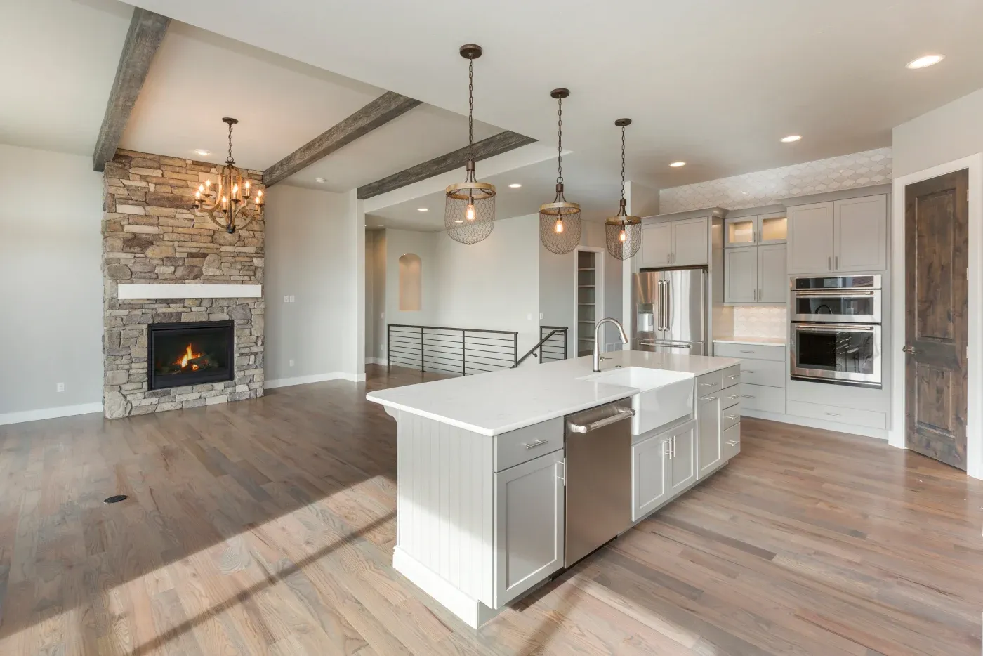 Modern kitchen with a large island, stainless steel appliances, pendant lights, wood flooring, and a stone fireplace in an open-concept living area.