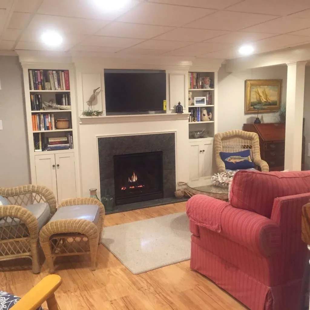 Cozy living room with a lit fireplace, surrounded by bookshelves. Wicker chairs and a red armchair create a warm, inviting atmosphere.