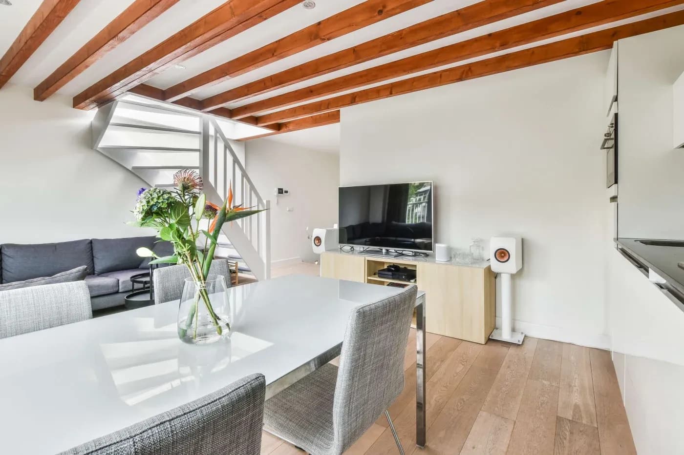 A modern living room with wooden floors, white walls, and exposed beams. It features a glass table with flowers, gray chairs, a sofa, a staircase, and a TV.