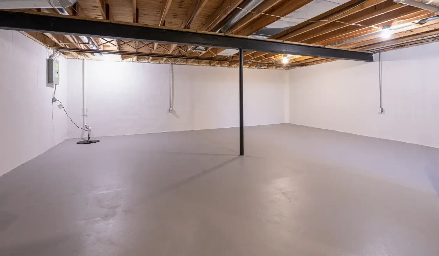 Spacious empty basement with white walls and a smooth gray concrete floor. Exposed wooden ceiling beams and minimal lighting create a neutral tone.