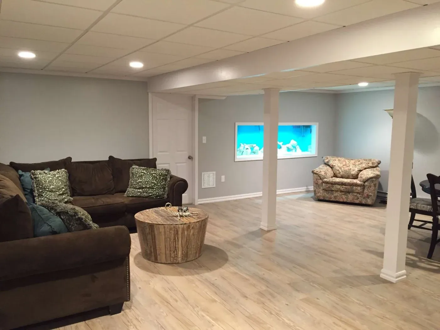 Modern basement with light wood floors, brown sectional sofa, and round wooden coffee table. A wall-mounted aquarium adds a calming touch.