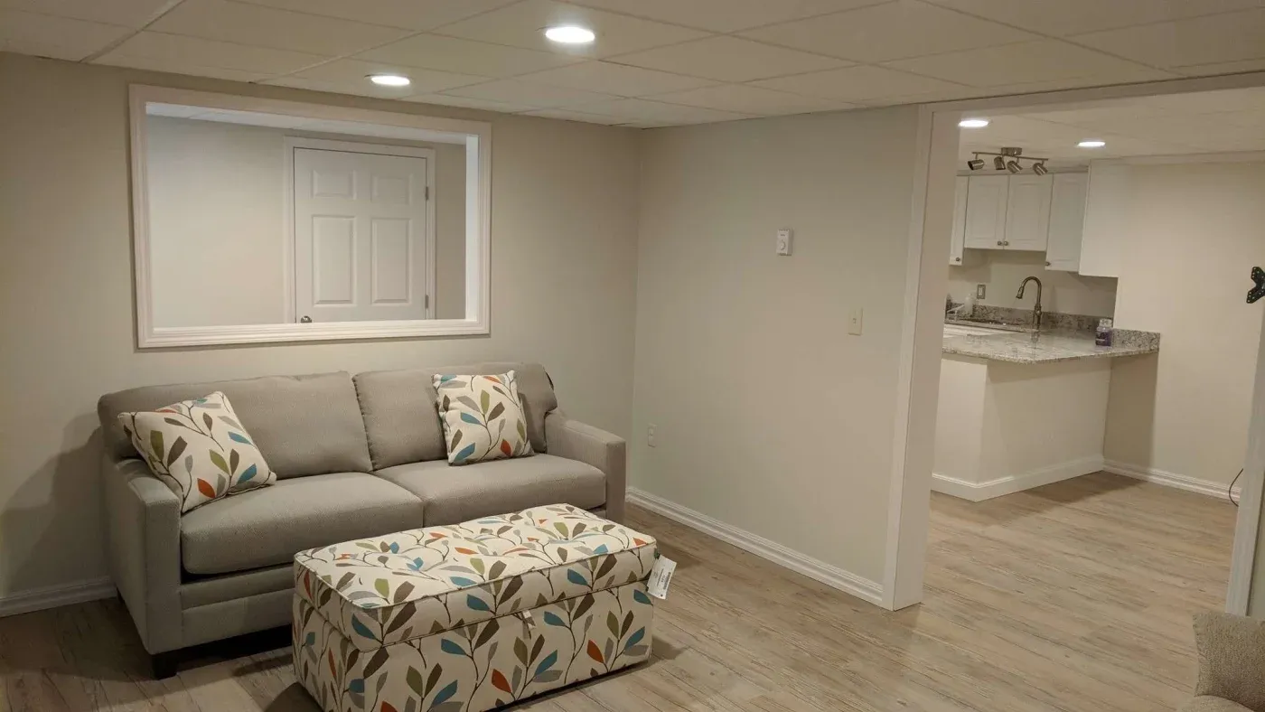 A cozy basement living area featuring a gray sofa with leaf-patterned pillows, a matching ottoman, light wood flooring, and a glimpse of a modern kitchen.