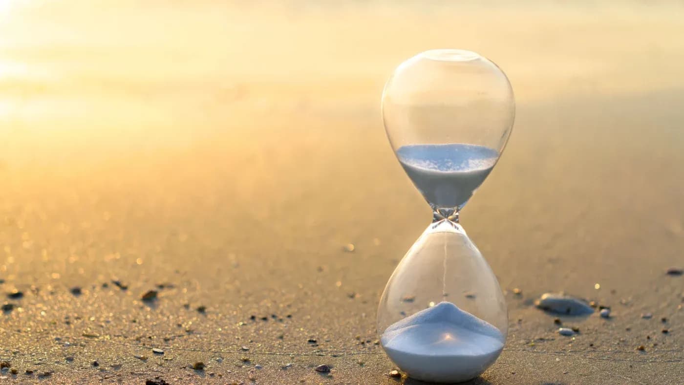 An hourglass with white sand stands on a serene beach at sunrise. Soft golden light reflects off the sand, conveying a peaceful, contemplative mood.