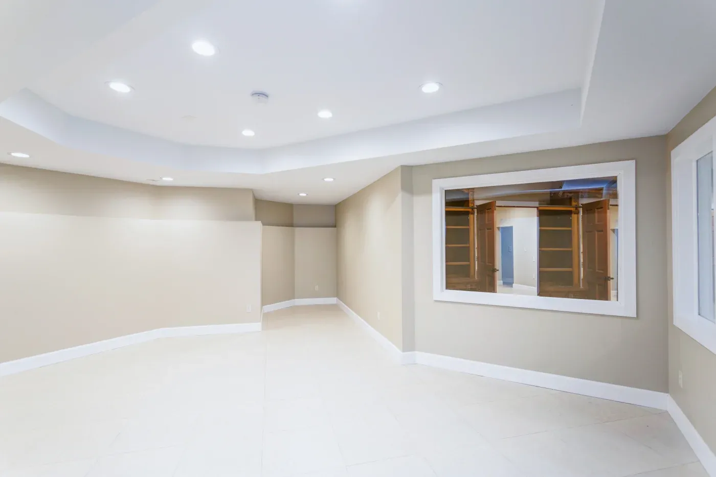 A spacious, empty room with beige walls and white tile flooring. Recessed ceiling lights illuminate the room. A large window reveals built-in wooden shelves.