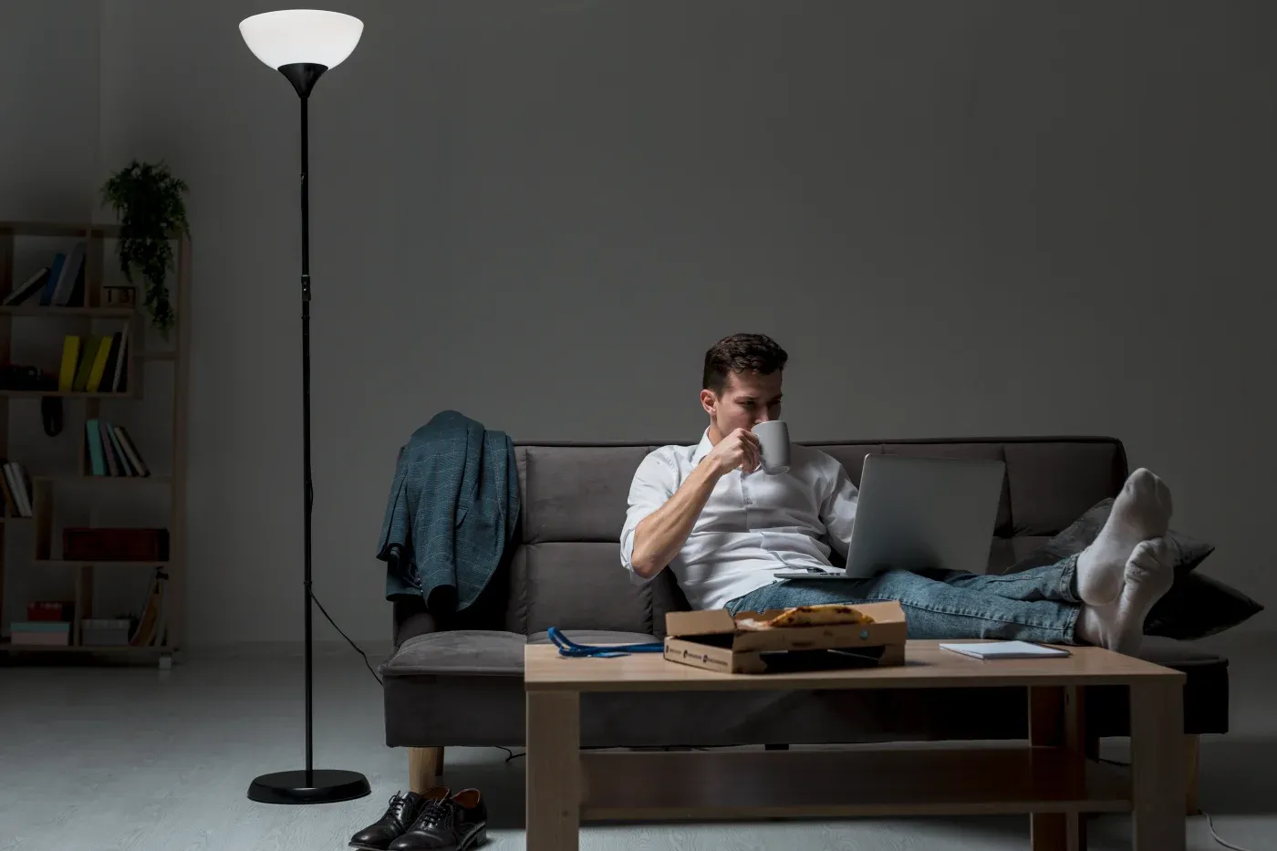 Man relaxing on a couch, using a laptop with feet up on a coffee table. Dimly lit room with floor lamp, books, a pizza box, and casual shoes nearby.