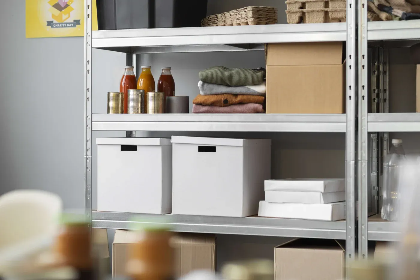 Metal shelves in a storage room hold canned goods, bottled sauces, folded clothes, and cardboard boxes, conveying organization and preparation.