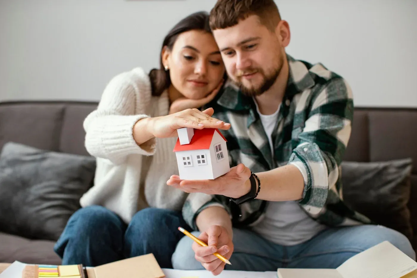 A couple sits close together on a couch, holding a small house model. They appear focused and thoughtful, with notebooks and pencils on the table.