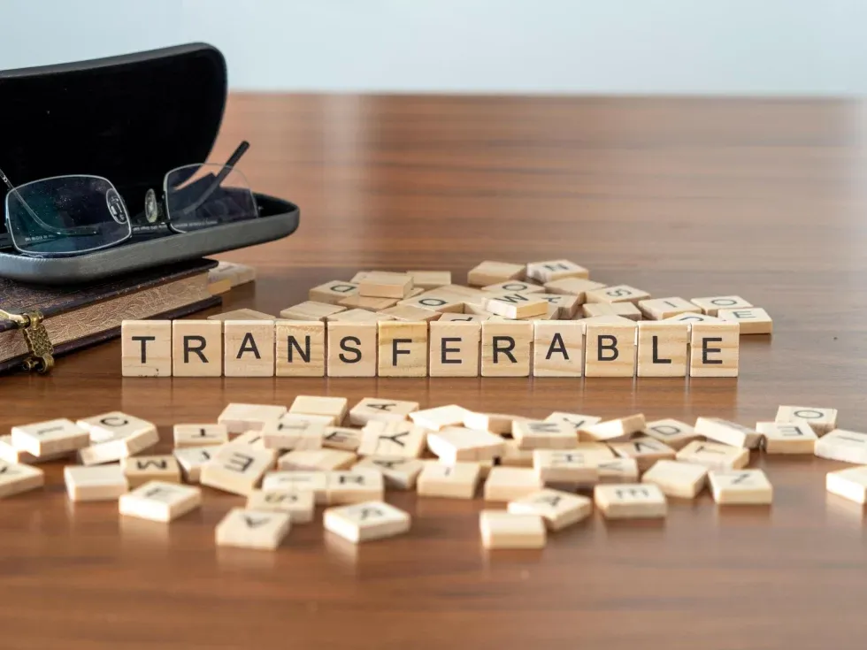 Wooden Scrabble tiles spell "TRANSFERABLE" on a table, surrounded by scattered tiles. Nearby are an open glasses case and a closed notebook.