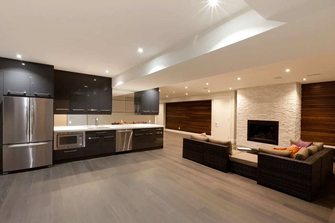 Modern open-plan kitchen and living room with sleek, dark cabinets, stainless steel appliances, a cozy sofa set, and a stone fireplace. Warm lighting.