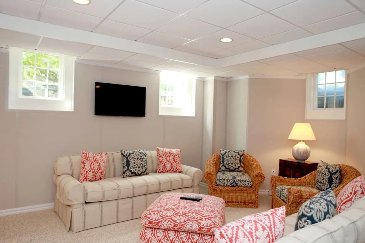 Cozy basement room with beige walls, two sofas, patterned cushions, a wicker chair, and a lamp. A small TV is mounted on the wall. Bright and inviting.